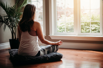 Woman sitting on the floor indoors meditating in front of windows.