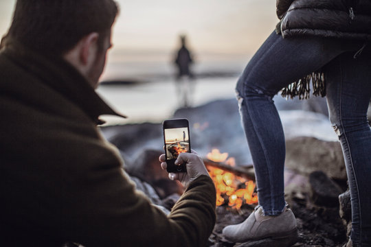 A man takes a picture of a campfire with his cell phone or smartphone