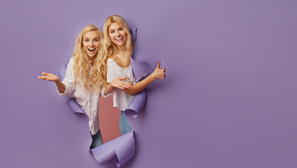 Surprised young women stands in torn paper hole, has excited cheerful expression, looks through...