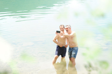 Two men standing together outdoors taking a selfie and smiling. Best friends taking a selfie while standing into the water of the lake on a summer day. Men wearing only jeans shorts. Bare-chested .