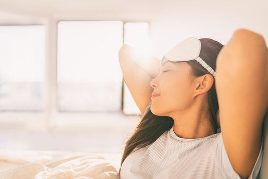 Sleeping At Home Woman Waking Up In Morning Sunlight Stretching Happy After A Good Night Sleep Feeling Rested Wearing Eye Mask. Asian Girl Bed Sleep In Hotel Bedroom Healthy Lifestyle.