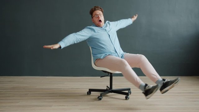 Happy young businessman in casual clothing is spinning on office chair indoors having fun on black background. Positive emotions, happiness and interior concept.