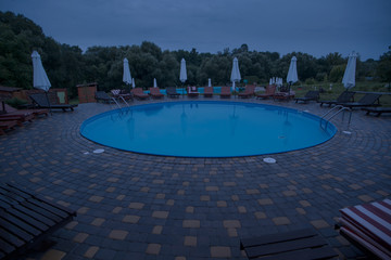 The round swimming pool with wooden deckchairs and umbrellas at night..