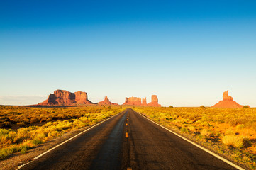 Empty straight road disappearing over the horizon dotted with iconic sandstone mesas of America's...