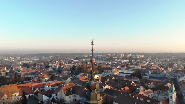 Drone fly around a bronze tower bell with all the baroque town above, smoke from factory during the sunset.