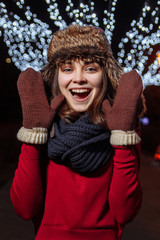 Satisfied charming woman wearing knitted scarf and mittens standing on street with illuminated garlands and expressing joy while looking at camera on Christmas 