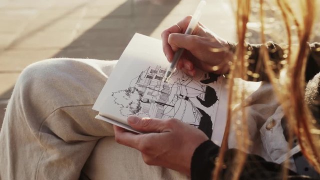 The artist makes sketches on the sunny promenade. Girl draws a coastal landscape with fishing boats. Black and white drawing by liner on white paper. Golden sunlight. Drawing outdoors. Hand drawing.