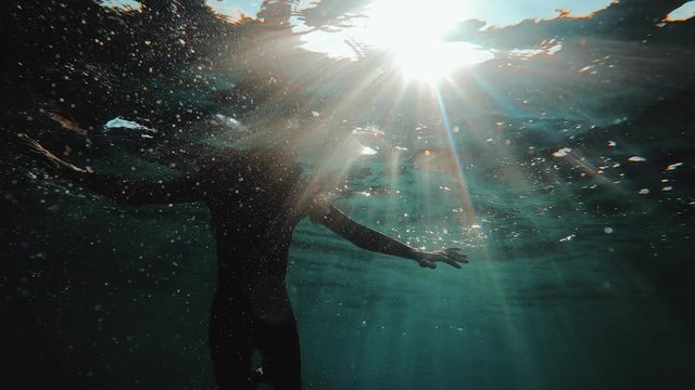 Redhead girl in a black neoprene suit splashes and swims underwater. Sun rays in clear emerald water. Spray and sun glare on the sea sand bottom. Swimming in open water. Beautiful underwater landscape