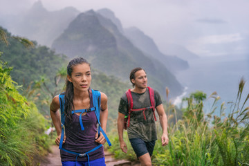 Hiking hikers woman and man trekking on trail trek with backpacks living healthy active lifestyle. Hiker girl walking on hike in mountain nature landscape wet scenery, Na Pali Coast, Kauai, Hawaii. - Powered by Adobe