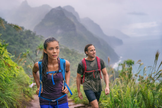 Hiking hikers woman and man trekking on trail trek with backpacks living healthy active lifestyle. Hiker girl walking on hike in mountain nature landscape wet scenery, Na Pali Coast, Kauai, Hawaii.