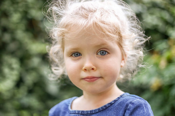 Portrait of a little beautiful European girl with curly blonde hair and blue eyes of 3 to 4 years in a classic blue color dress, trendy color 2020. Girl plays in the garden outside in spring or summer