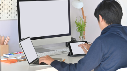 Back view of young creative man while using white blank screen laptop, tablet and computer at the working desk.