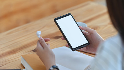 Cropped shot of young woman while holding earphone and white blank screen smartphone on wooden desk.