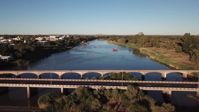 4K summer sunny afternoon aerial drone video: Orange River arched motorway long bridge, grassy islands, boat on the water and busy N10 highway located in town Upington, Northern Cape, South Africa
