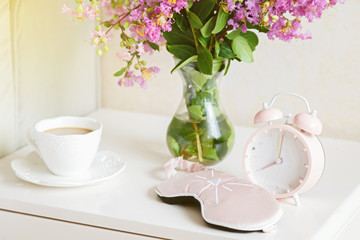 Alarm clock closeup, a cup of coffee and bouquet of pink flowers background in the morning sunlight