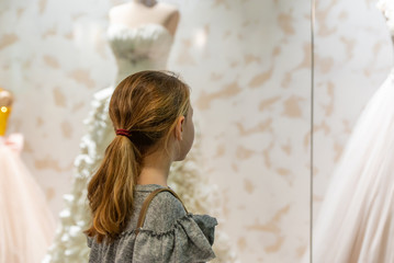 A young girl, a teenager, stands near the window of a wedding salon and looks at wedding dresses, introducing family life.
