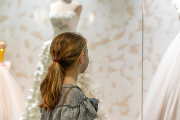 Fototapeta premium A young girl, a teenager, stands near the window of a wedding salon and looks at wedding dresses, introducing family life.