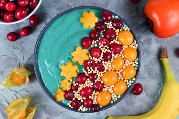 Top view of healthy yogurt and fruit smoothie bowl with natural green spirulina powder and decorated with cranberries, physalis and kaki fruits and puffed quinoa grains