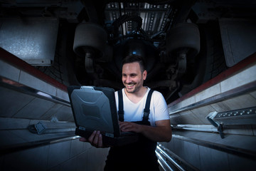 Vehicle mechanic with diagnostic tool laptop working under the truck in workshop. Experienced...