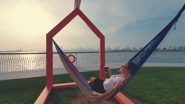 Young woman weared in office sute having coffee in hammock while chilling , freelance life style conceptual, work anywhere. Modern city on the background.