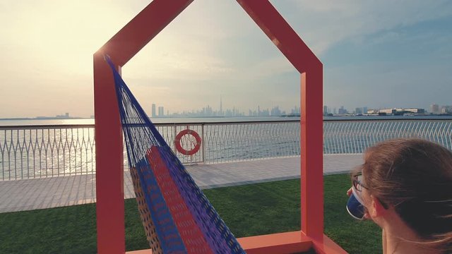 Young woman weared in office sute having coffee in hammock while chilling , freelance life style conceptual, work anywhere. Modern city on the background.