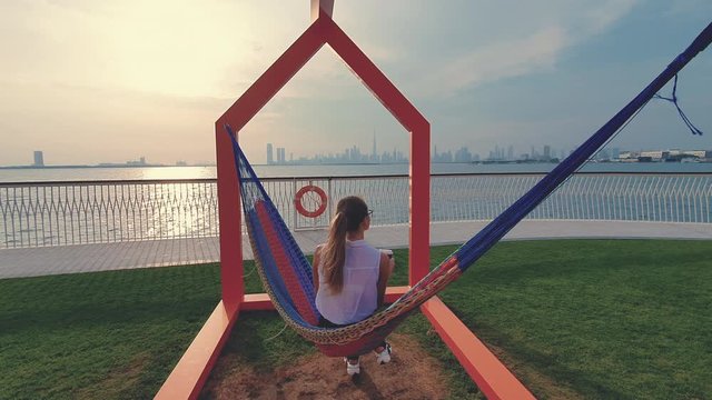 Young woman weared in office sute having coffee in hammock while chilling , freelance life style conceptual, work anywhere. Modern city on the background.