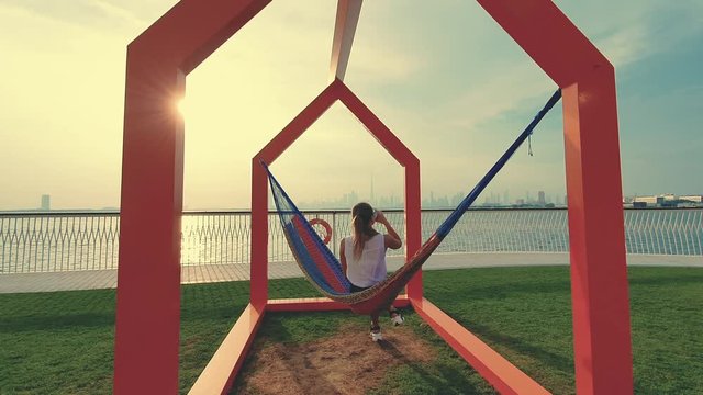 Young woman weared in office sute having coffee in hammock while chilling , freelance life style conceptual, work anywhere. Modern city on the background.
