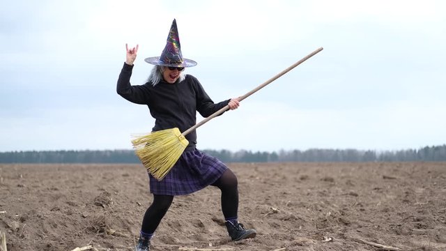 Funny woman in modern witch costume with cone hat, grey wig and dark glasses playing on broom   like on guitar and dancing on autumn empty field. Halloween concept.