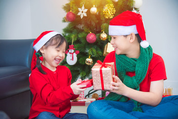 Fototapeta na wymiar Little asian girl and her brother sitting on the floor and giving gift box to each other with smiling together at home in Christmas holiday.