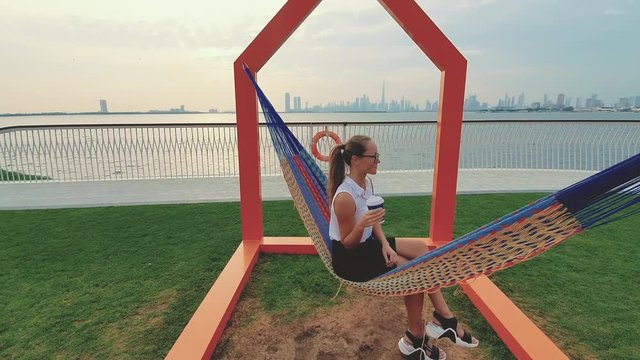 Young woman weared in office sute having coffee in hammock while chilling , freelance life style conceptual, work anywhere. Modern city on the background.