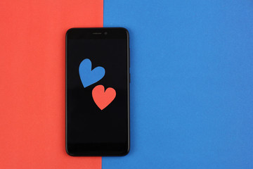 black smartphone and two craft paper valentine hearts red and blue on color papers background, copy space on blue color