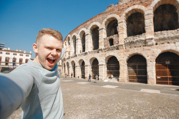 Happy male traveler makes selfie photo on background of amphitheater coliseum in city Verona Italy. Concept travel