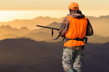 Tuinposter A man with a gun in his hands and an orange vest on a pheasant hunt in a wooded area in cloudy weather. Hunter with dogs in search of game. © Mountains Hunter