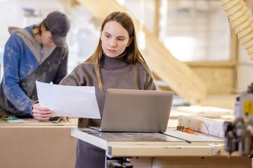 Charming young concentrated girl designer designs sketches of new collection of furniture on wooden production. Concept of successful career as an engineer or manager. Place for text