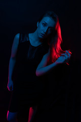 Young girl in a black dress. Girl playfully leaned forward. Bright red hair from the light In the light of colored lamps of blue red colors. Neon lights effect lamps. on a black background