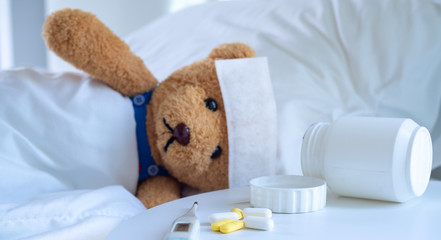 The teddy bear and the fever-reducing patch lie on the bed in a white-tone bed next to a medicine...