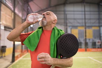 Mature man in paddle tennis court with towel and bottle of water.