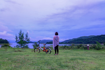 A woman standing at big lake and mountain range on holiday near a bicycle 