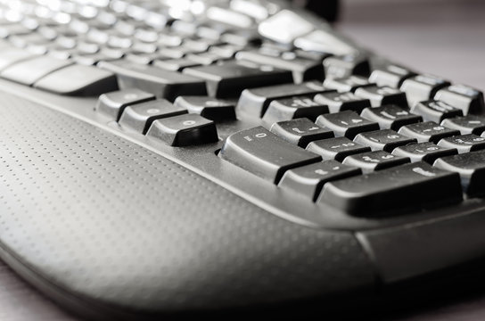 Close-up Of A Dark Dusted Computer Keyboard. Black Ergonomic Keyboard On A Light Gray Desk. Eye Level Shooting. Selective Focus.