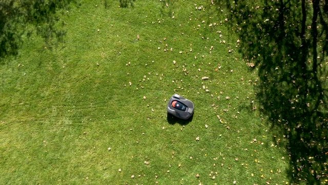 Robotic lawn mower moving across lawn from aerial view while rising high into the sky from drone.