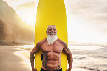 Happy fit senior having fun surfing at sunset time - Sporty bearded man training with surfboard on the beach - Elderly healthy people lifestyle and extreme sport concept - obrazy, fototapety, plakaty