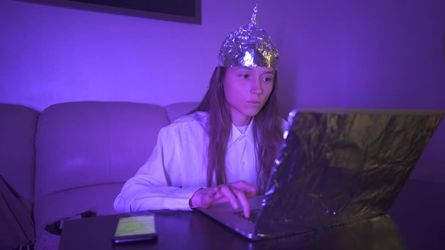 Young woman in Tin Foil Hat typing text on keyboard, laptop with foil, then take mobile phone. Foil hat shields her from 5G waves, electromagnetic fields, mind control, mind reading, global conspiracy