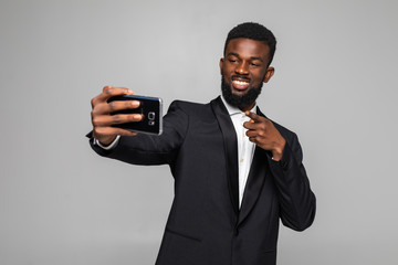 Happy young handsome African businessman taking selfie pointed on camera isolated on gray background