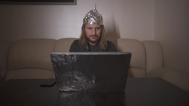Exhausted man put on aluminium foil hat on head and search in internet information about conspiracy, type text on laptop. Foil hat shields him from 5G waves, mind control, mind reading, illuminati.