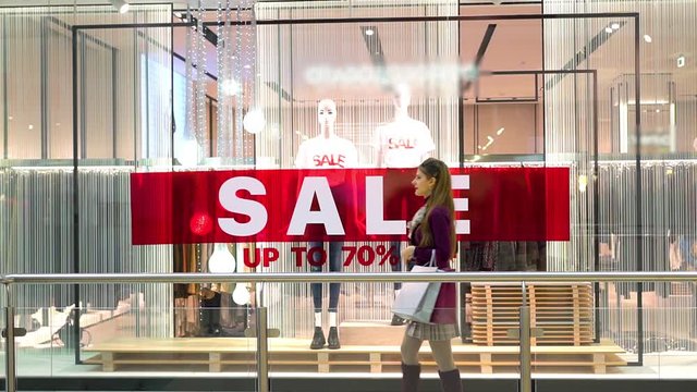 A slim, elegant woman shopper walks with bags past a huge red sale sign in the window of a fashion boutique in the Mall.