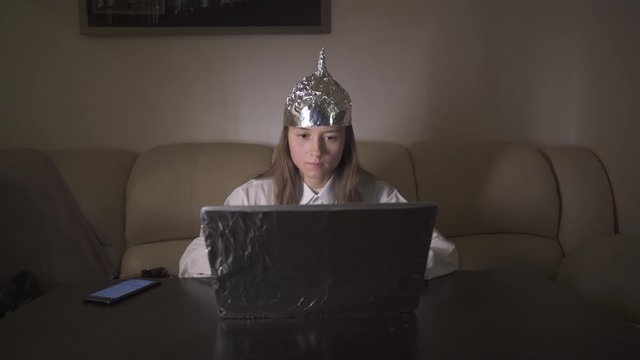 Woman in tin foil hat puts down phone and works at computer, typing on keyboard. Foil hat shields her from 5G waves, internet, electromagnetic fields, mind control and mind reading.