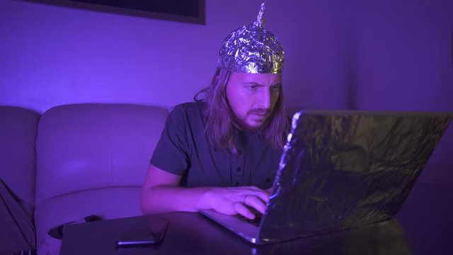 Man with long hair in Tin Foil Hat type message on phone, work at laptop. Man tired of computer, rubbing eyes. Foil hat shields him from 5G waves, electromagnetic fields, mind control, mind reading.