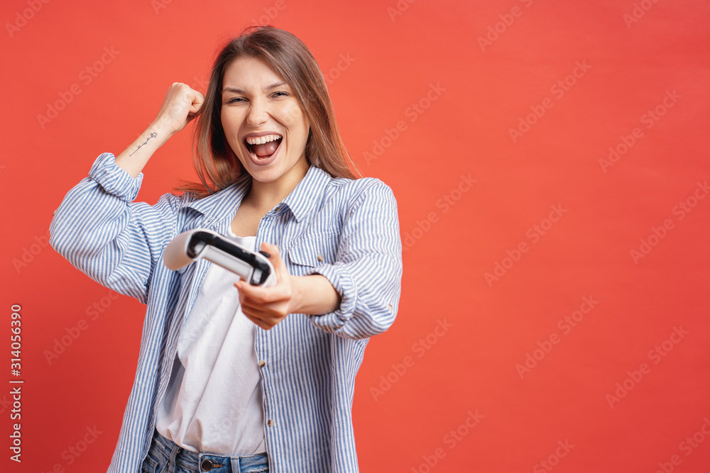 Wall mural competitive girl celebrates winning holding game joystick controller on red background.