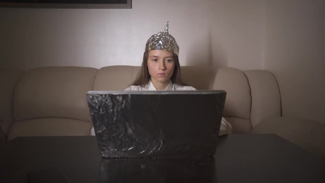 Exhausted woman put on aluminium foil hat on head and search in internet information about conspiracy, type text on laptop. Foil hat shields her from 5G waves, electromagnetic fields, mind reading.
