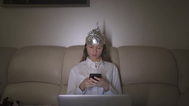 Tin Foil Hat protects woman`s brain from 5G waves, electromagnetic fields, mind control. Girl typing message in mobile phone.